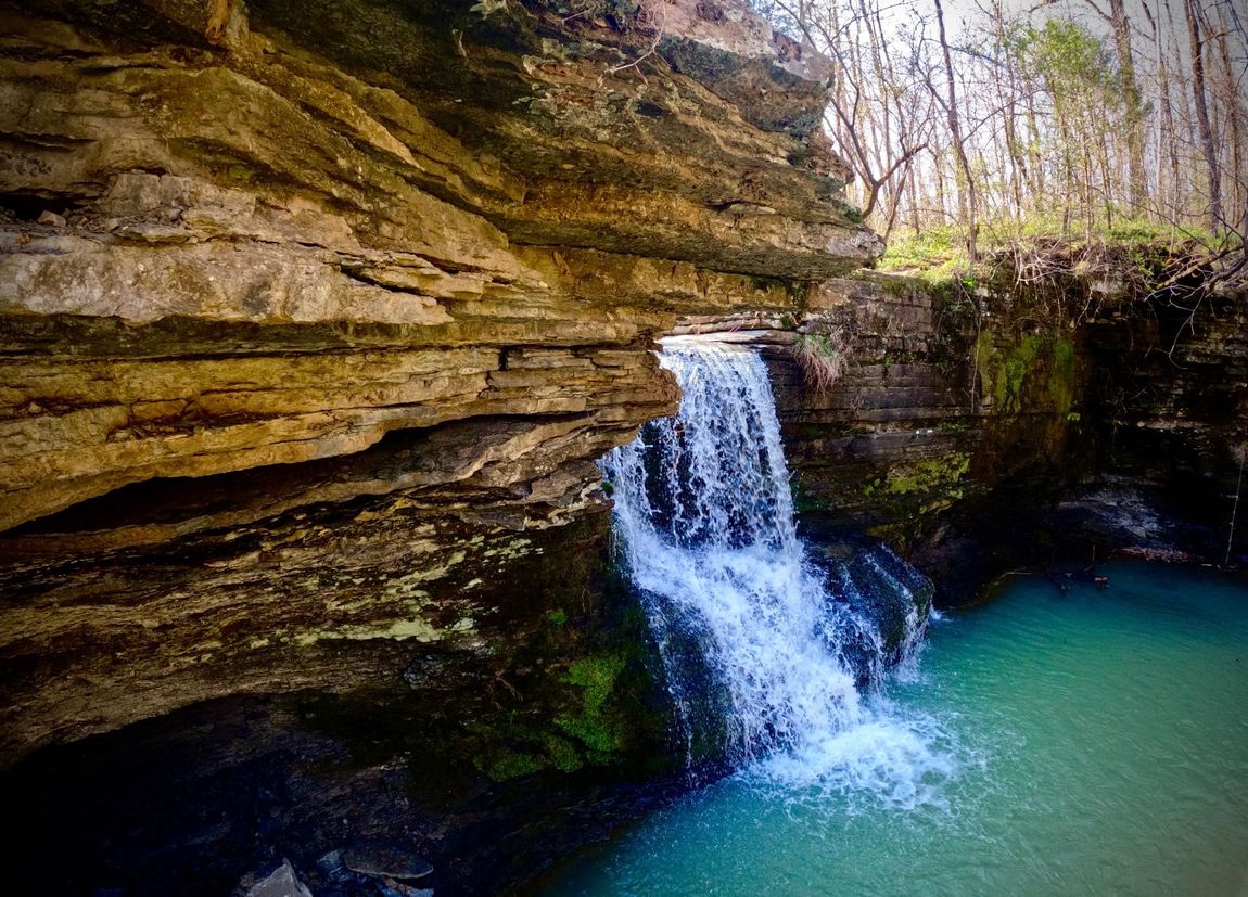 Small waterfall pouring over layered rock into a turquoise pool in a wooded canyon