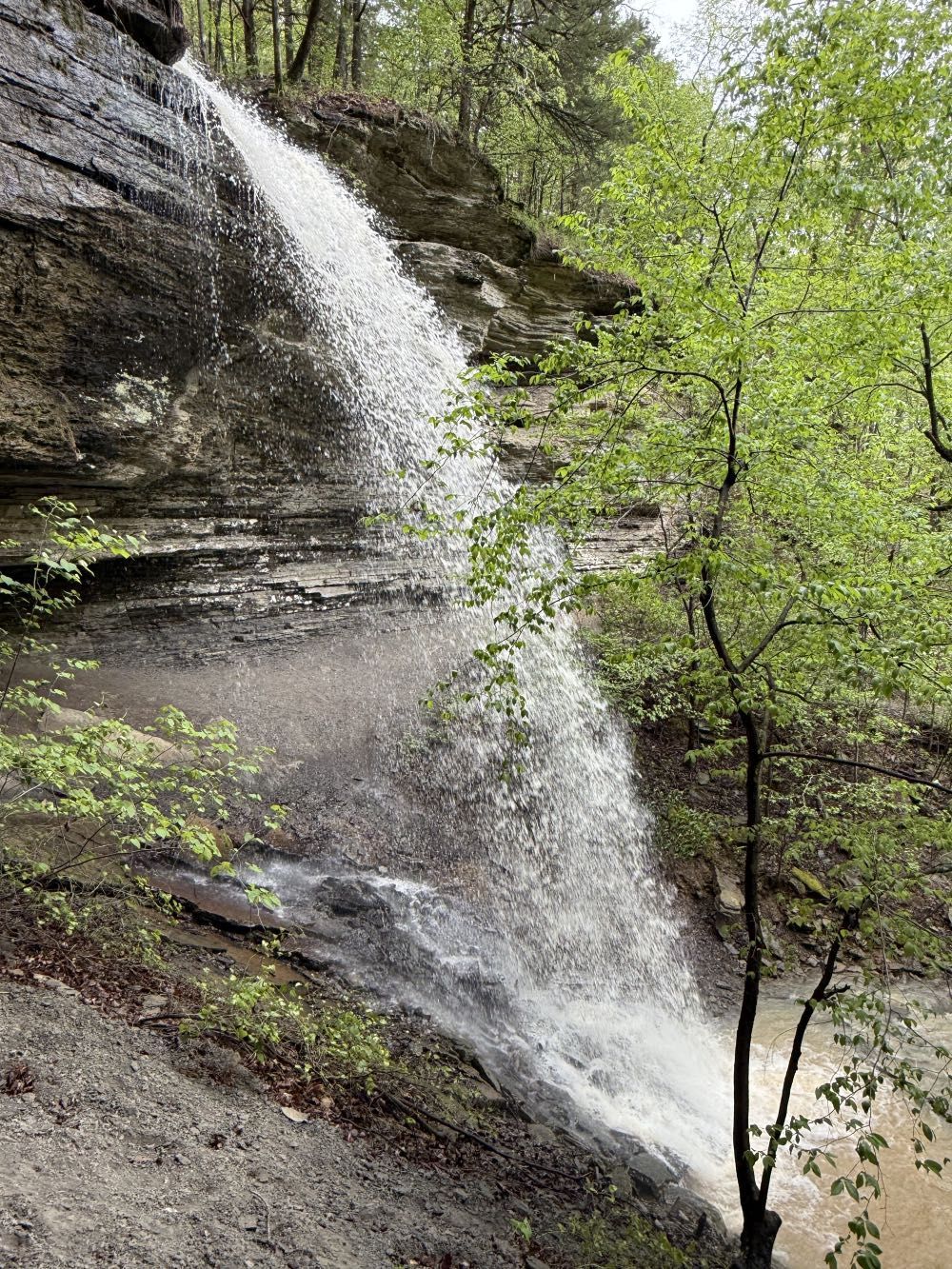 A camouflage off-road vehicle parked on a rocky ledge directly above a small waterfall in a forest.