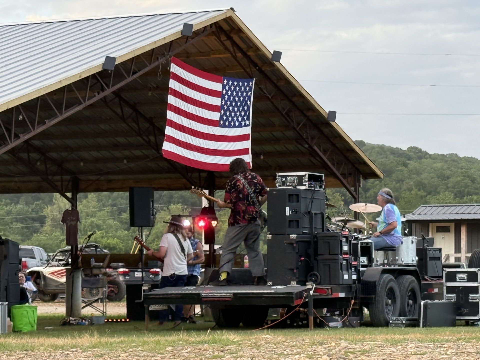 Outdoor stage with musicians performing under a large American flag at a rural pavilion