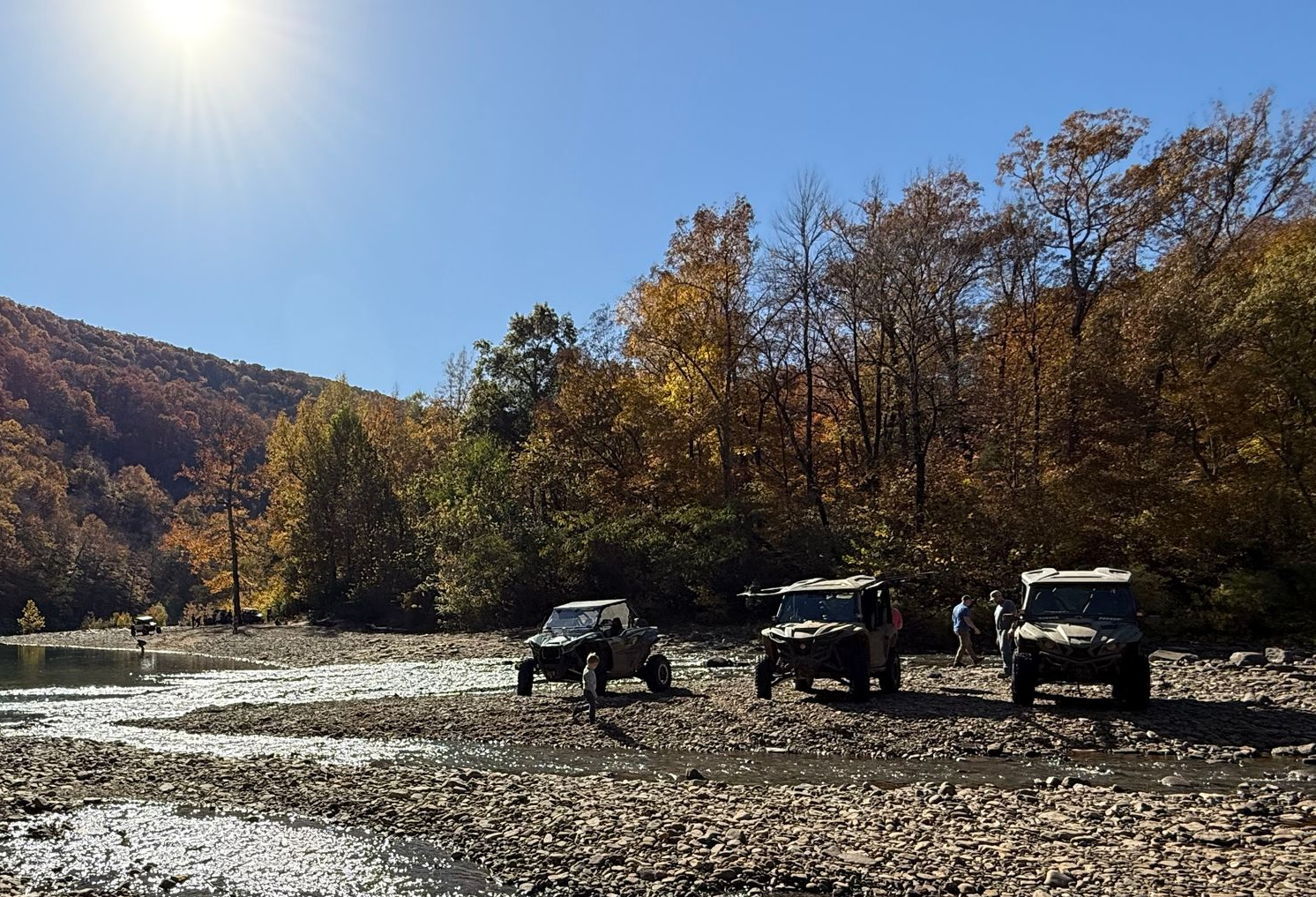 Three off-road vehicles parked on a rocky riverbed with autumn trees and hills under a clear blue sky