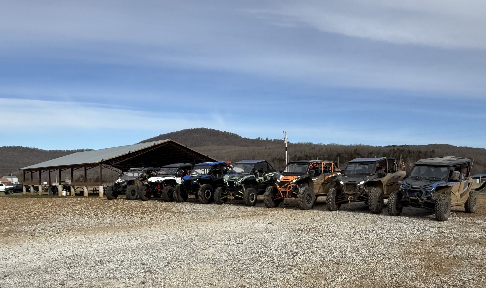 Line of off-road vehicles parked beside a barn on a gravel lot under a blue sky.