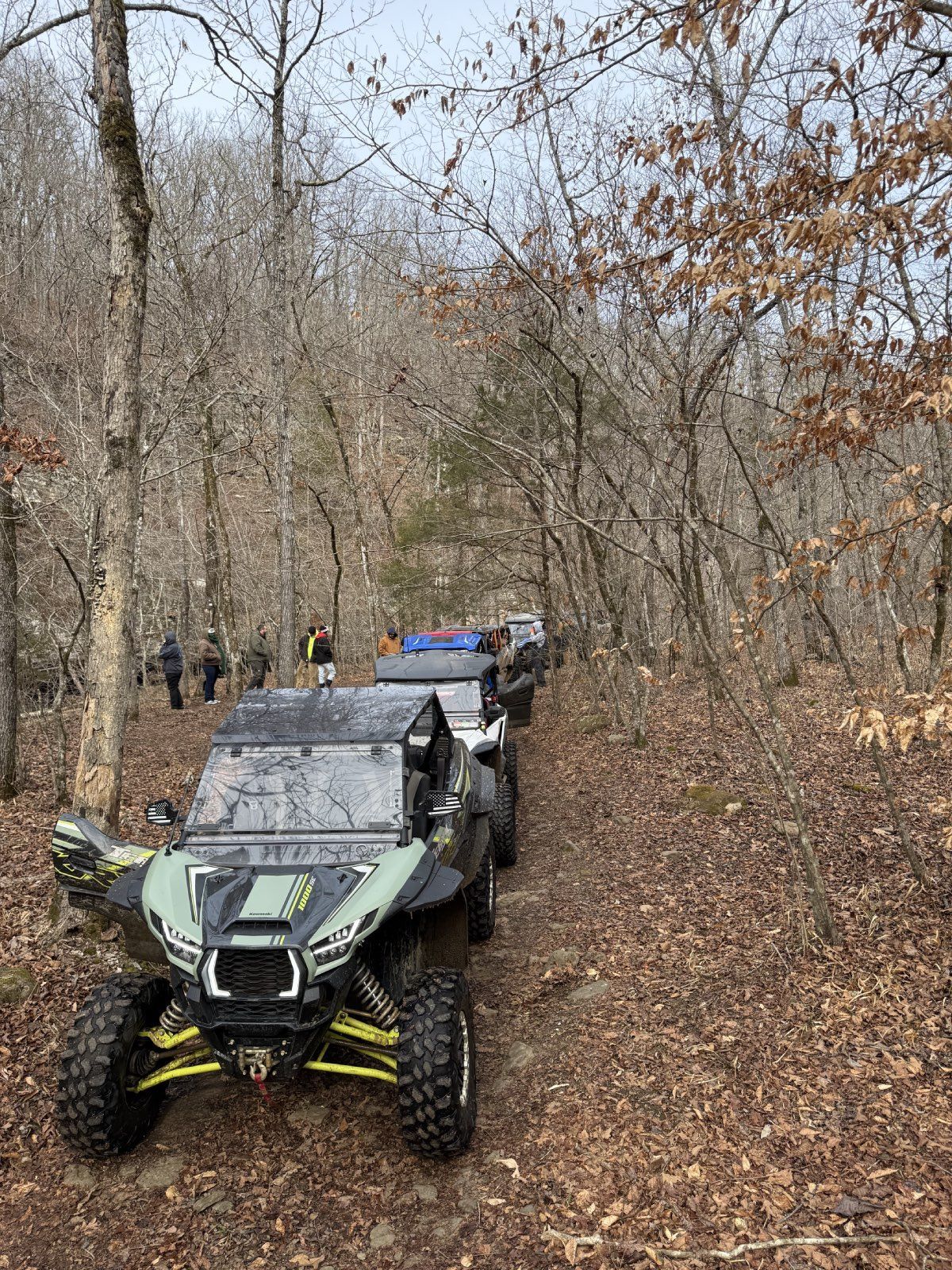 Off-road ATV with yellow suspension on a muddy forest trail in late autumn, with another vehicle behind it.