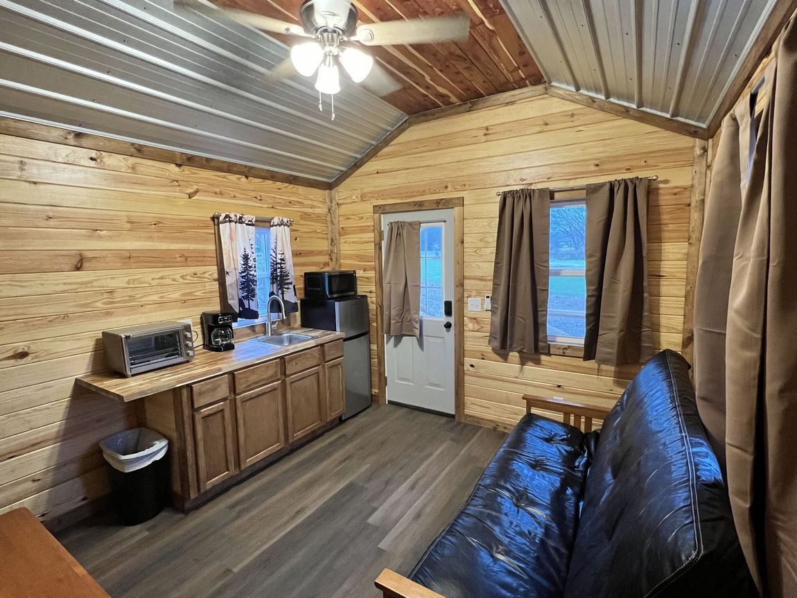 Cozy wood-paneled cabin interior with sofa, kitchenette, door, and windows with curtains.
