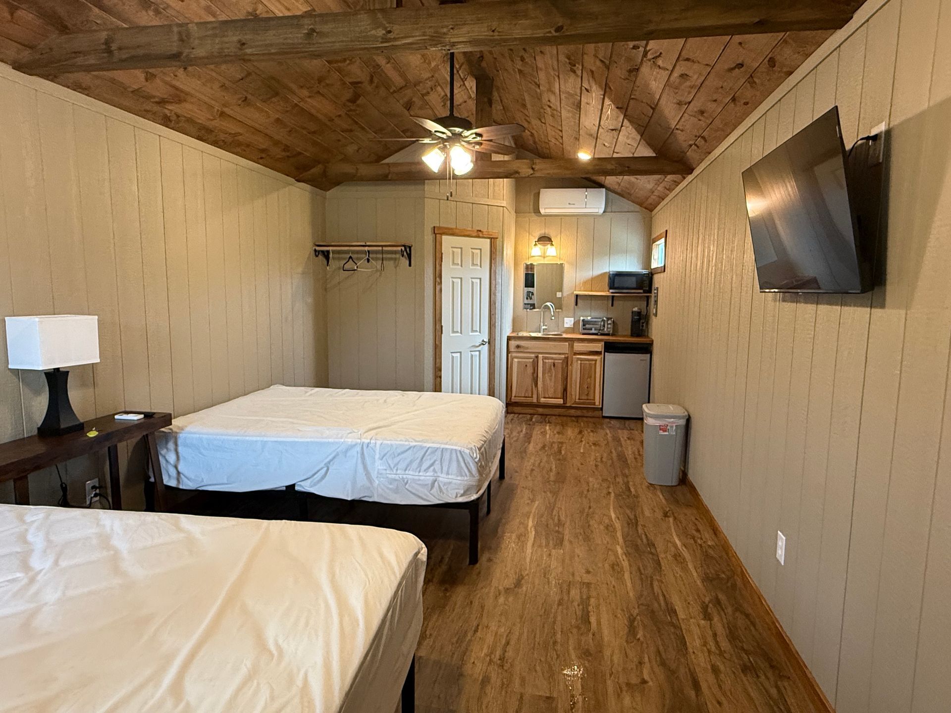 Small rustic bedroom with two beds, wood-paneled walls, ceiling fan, kitchenette, and wall-mounted TV.