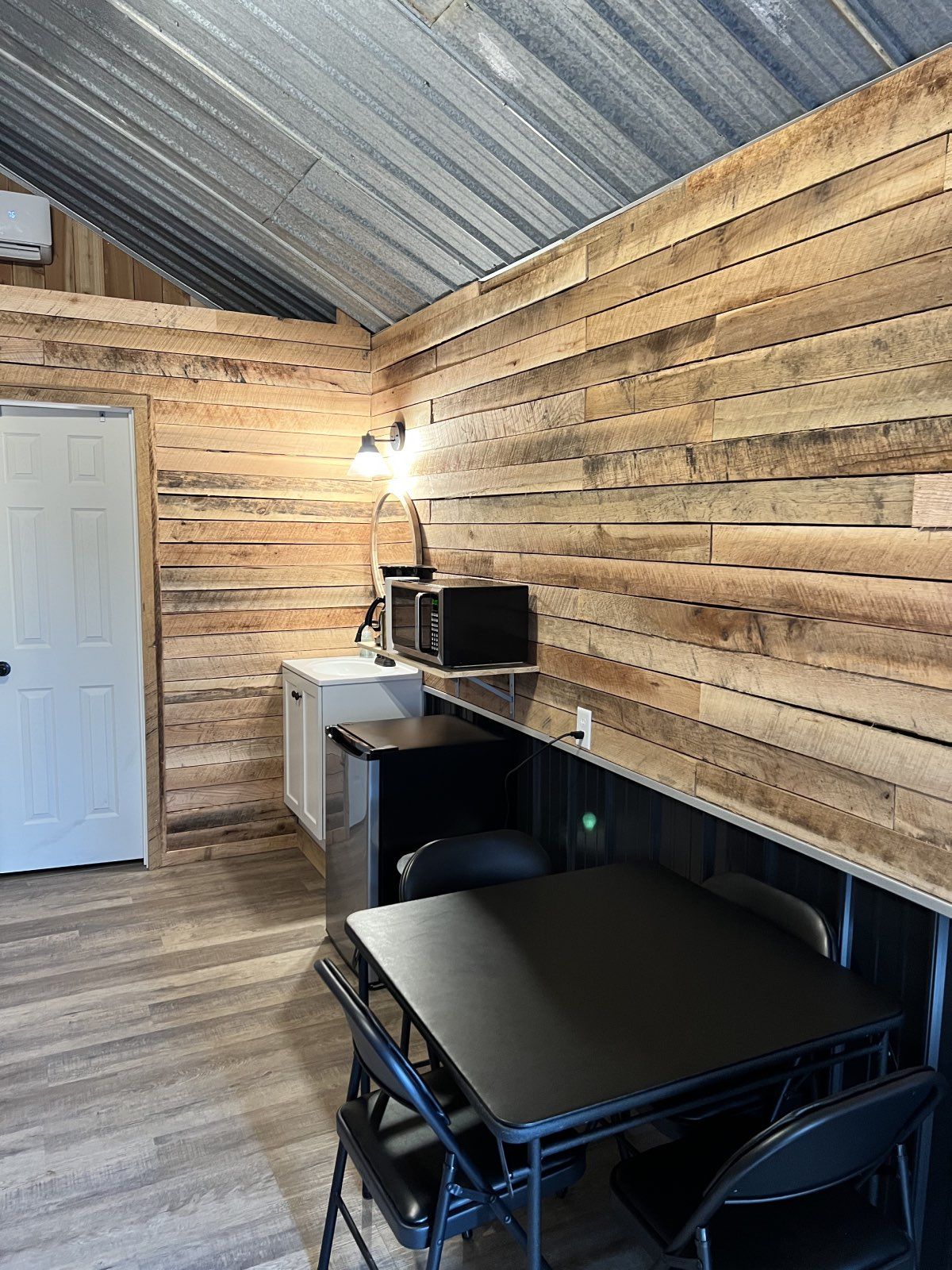 Rustic wood-paneled room with desk, black table, white door, and corrugated metal ceiling
