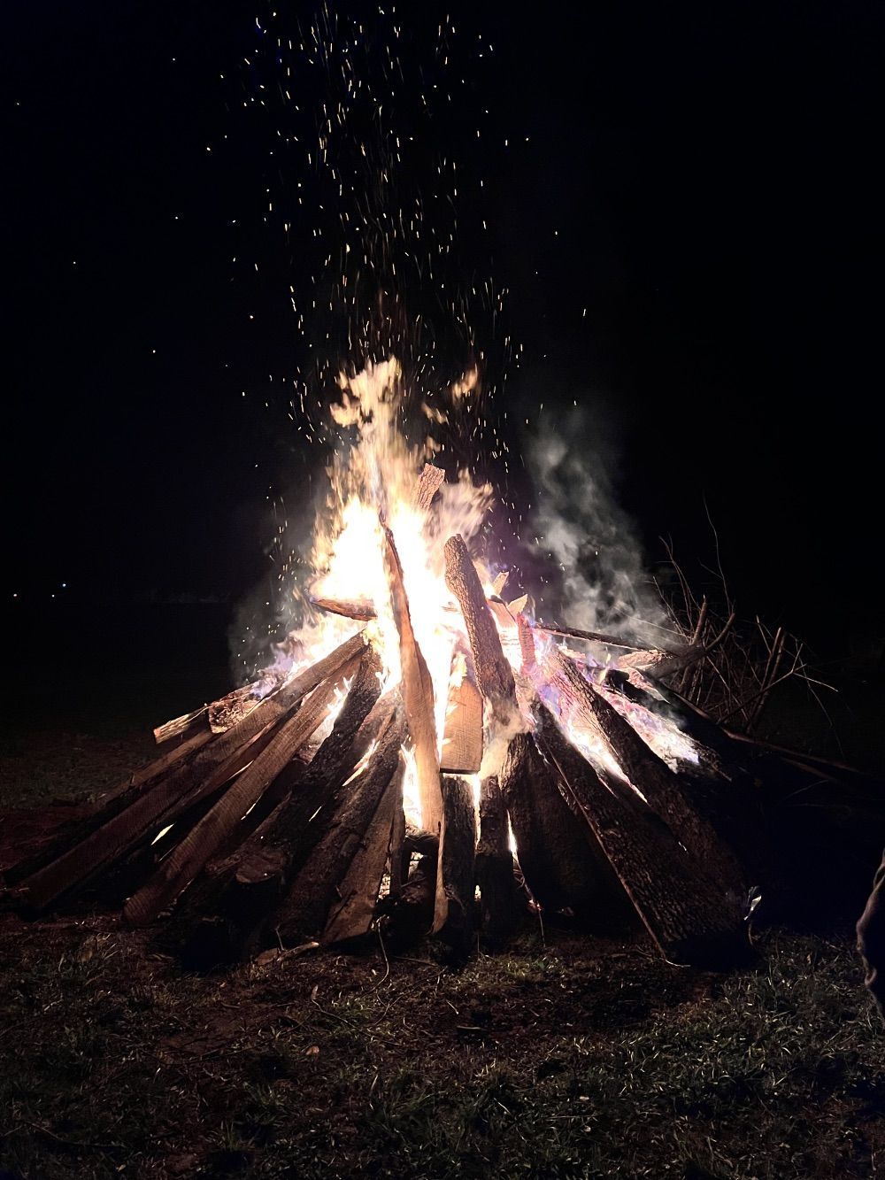 Bonfire burning at night, with sparks and smoke rising from stacked logs.