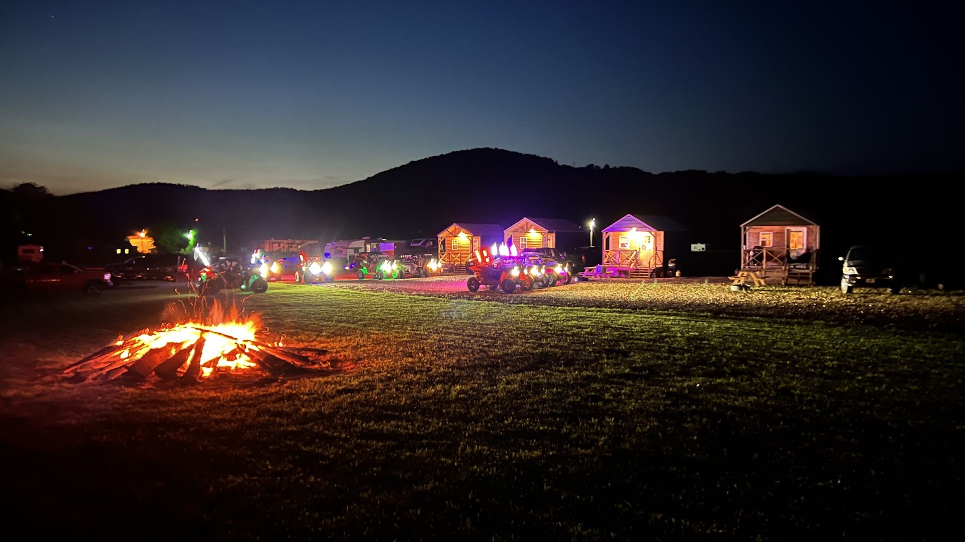 Night campsite with a bonfire, colorful lit tents, and cabins under dark hills