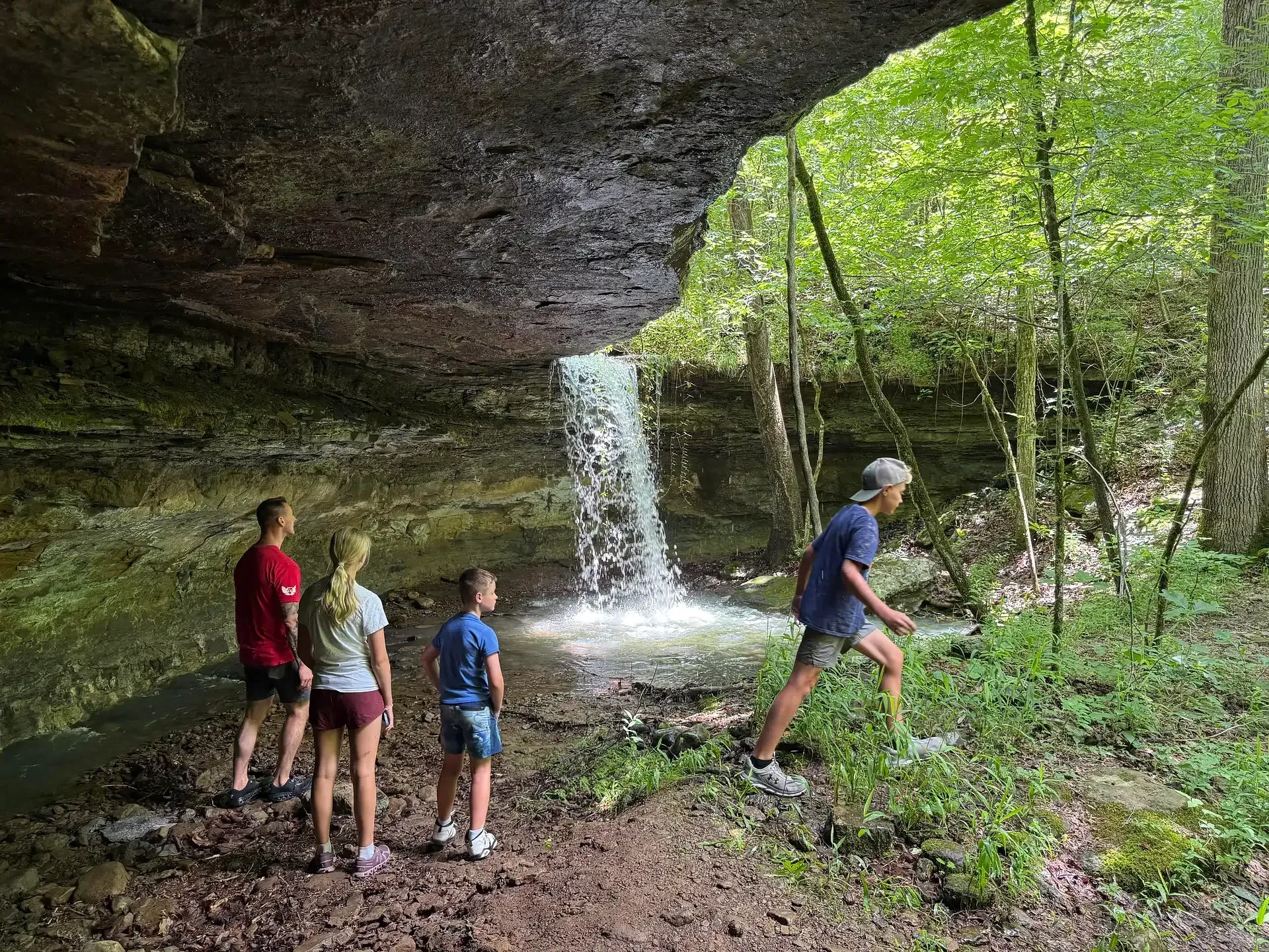 People hiking under a rock overhang near a small waterfall in a wooded area