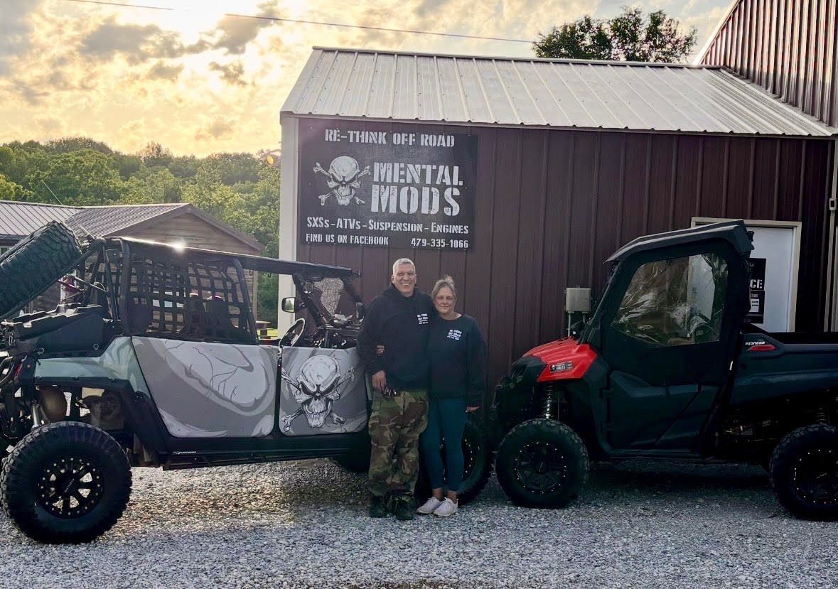 Two off-road vehicles parked by a barn, with three people standing between them at sunset.