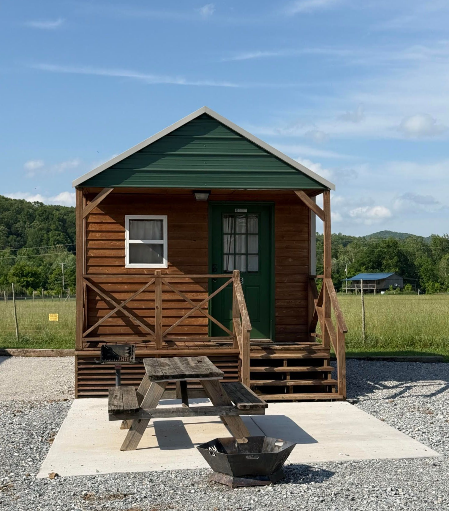 Small wooden cabin with porch and picnic table on a concrete pad in a grassy field under a blue sky