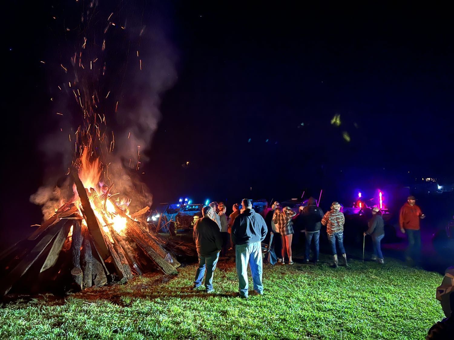 People gathered around a large bonfire at night, with fire trucks and flashing lights in the background.