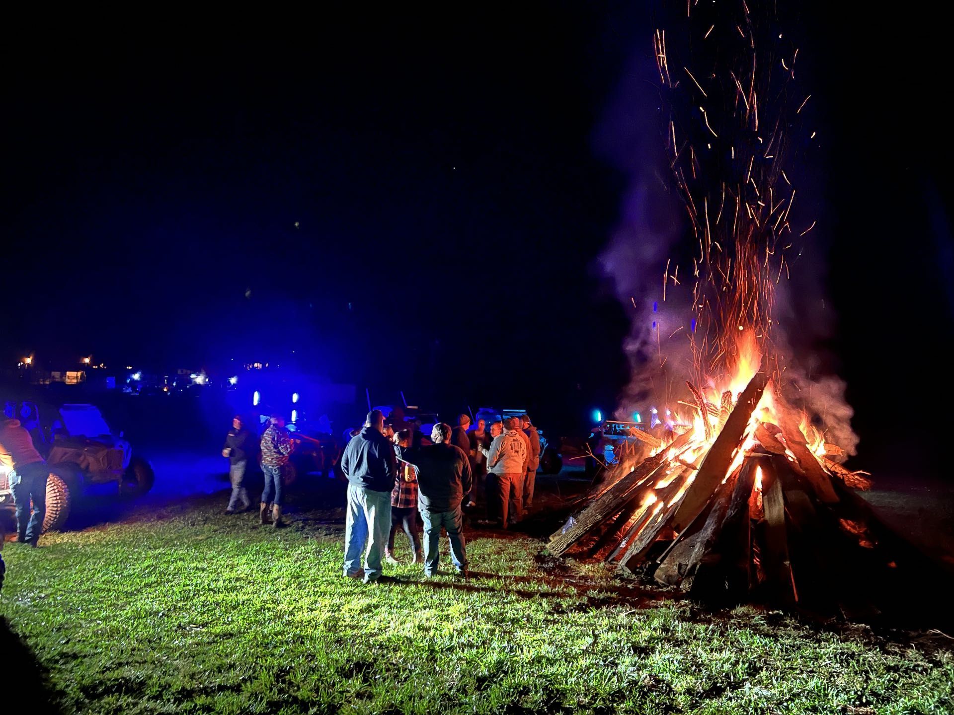 People gathered around a large bonfire at night, with sparks flying and blue lights in the background.
