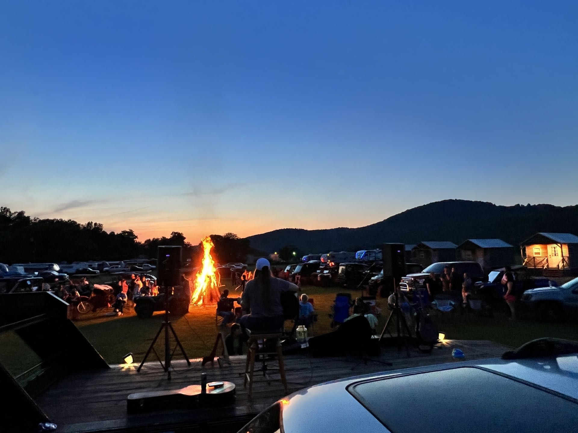 Crowd gathered around a bonfire at dusk in a campsite with mountains in the background