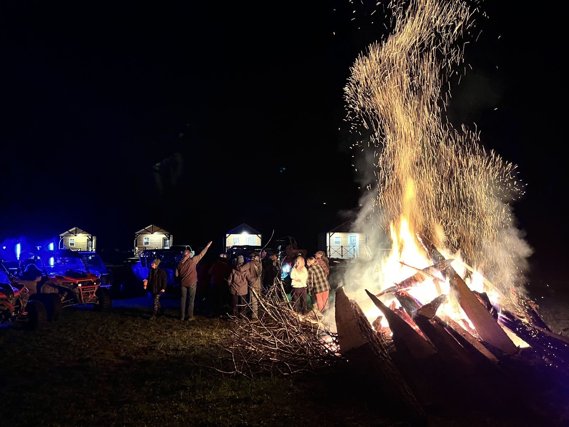 Bonfire blazing at night with a crowd gathered nearby under tents or lights