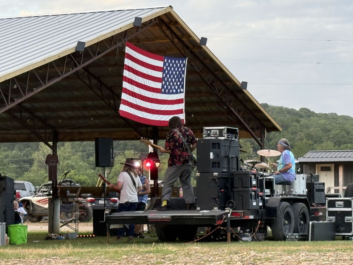 Outdoor stage with a large American flag and musicians performing under a pavilion