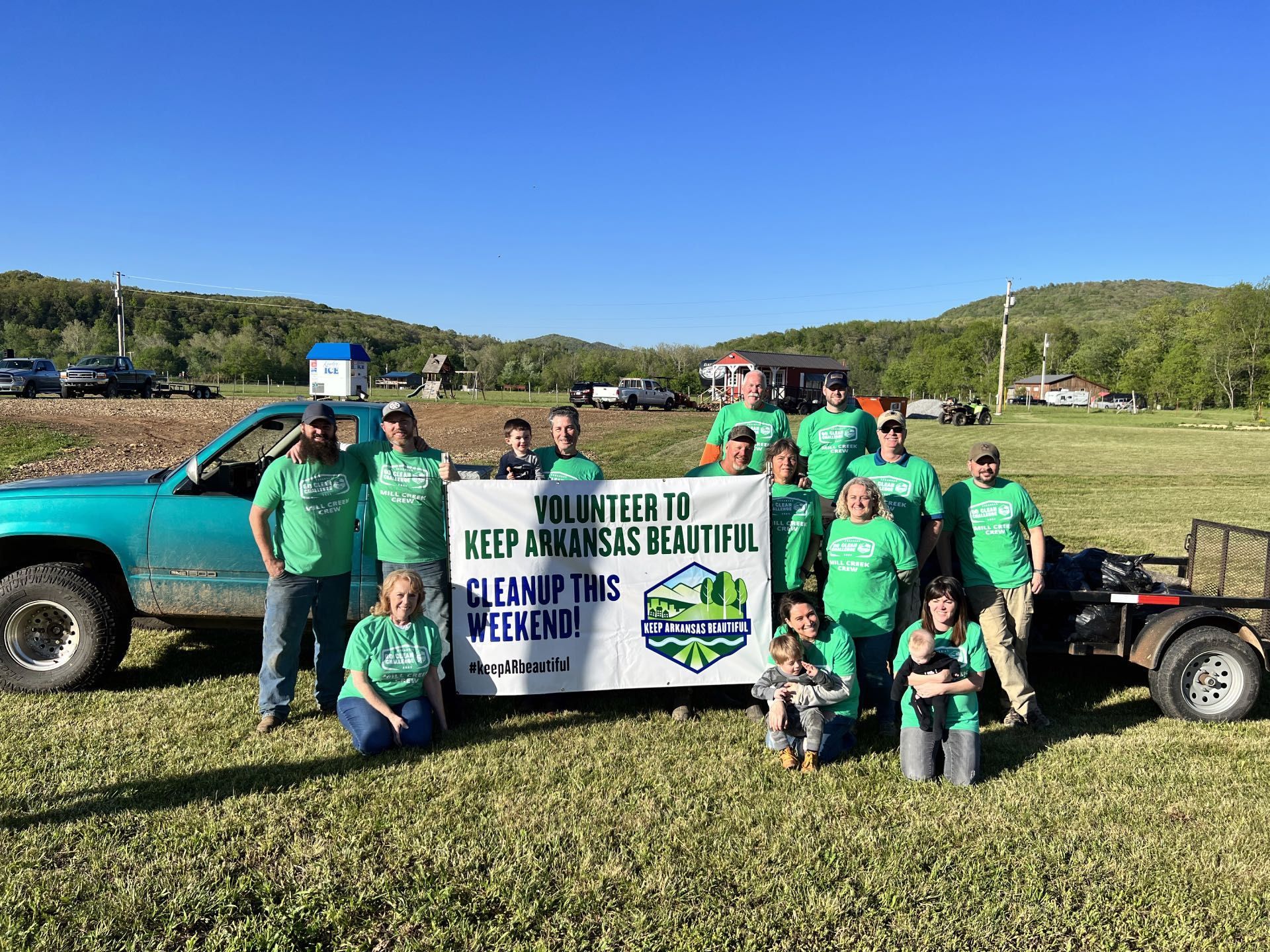 Group in green shirts holding a banner beside a turquoise truck and trailer in a grassy field, under a blue sky