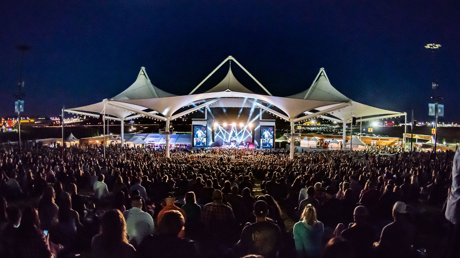 Night concert stage under tented canopy with a large crowd in front, lit by bright stage lights.