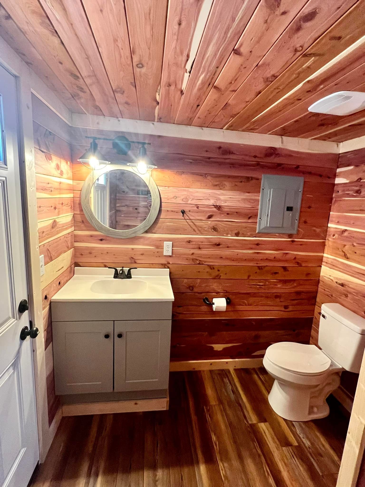 Small rustic bathroom with wood-paneled walls, white vanity, round mirror, and toilet.