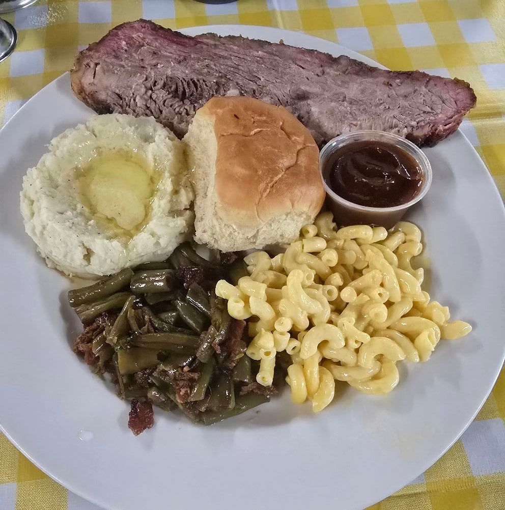 Plated barbecue meal with sliced meat, bread roll, cornbread, beans, green beans, macaroni, and sauce