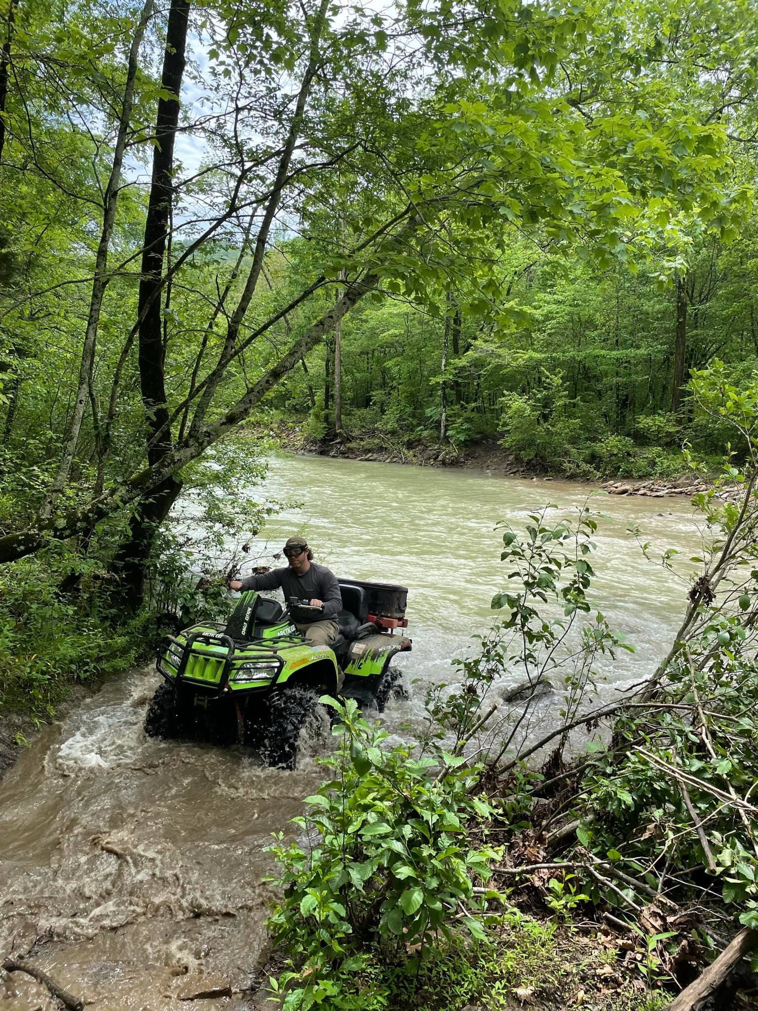 Person riding a green ATV through a muddy stream in a wooded area
