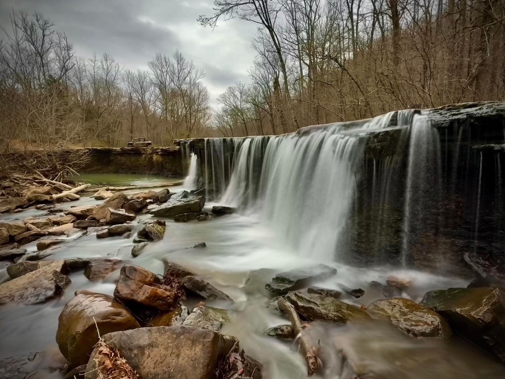 Waterfall cascading over a rocky ledge into a stream, surrounded by leafless trees under a cloudy sky
