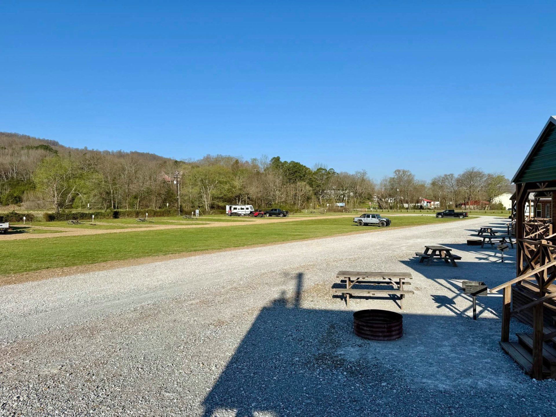 Gravel lot beside a rustic building with picnic tables, open field, trees, and a blue sky