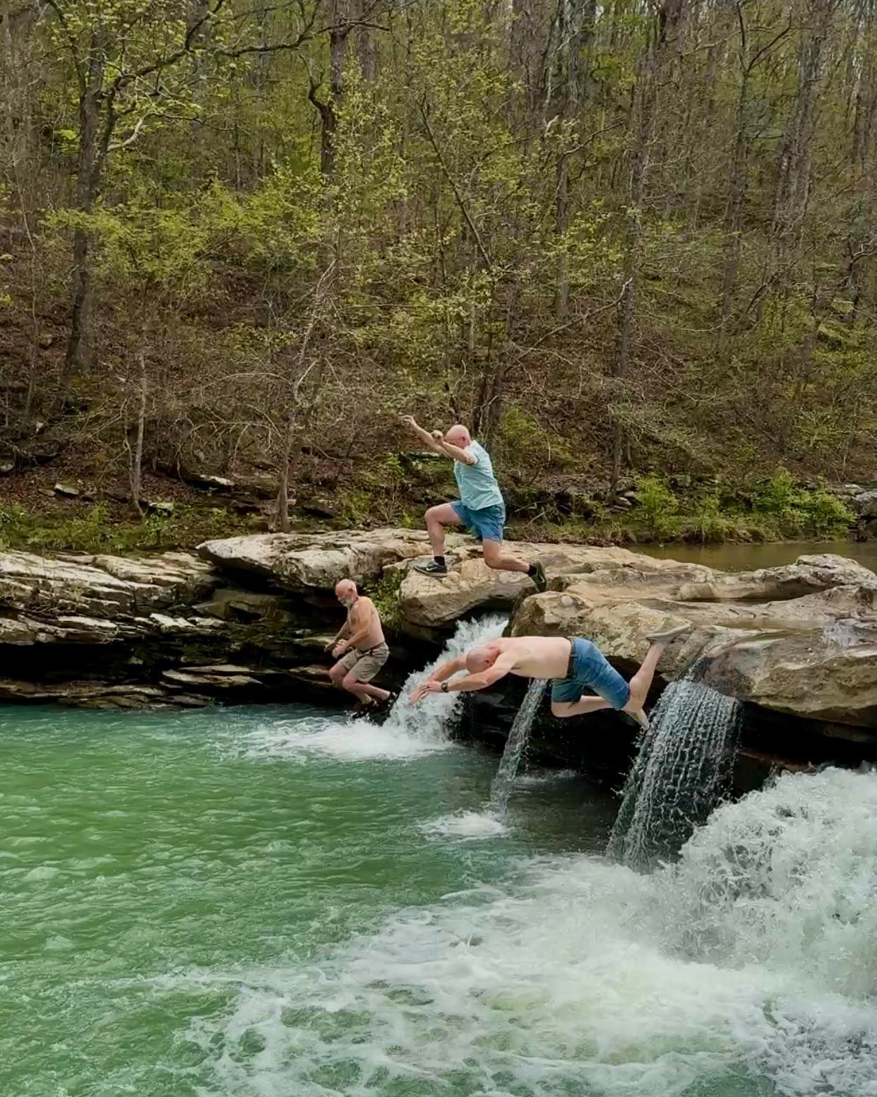 People swimming and jumping into a green river below a rocky ledge in a wooded area