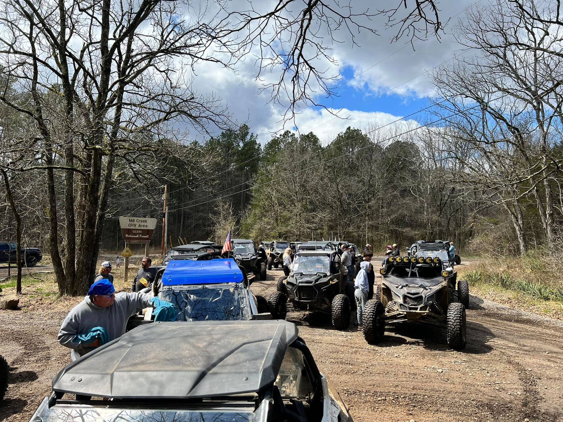 Off-road vehicles parked on a muddy trail in the woods under a partly cloudy sky.