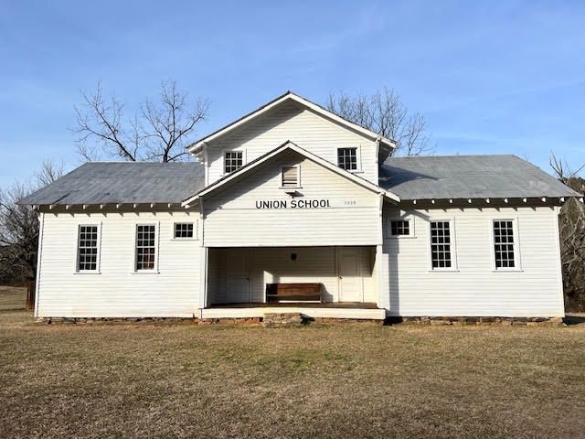 White schoolhouse with a central porch and sign reading “Union School” on a grassy lot under a blue sky