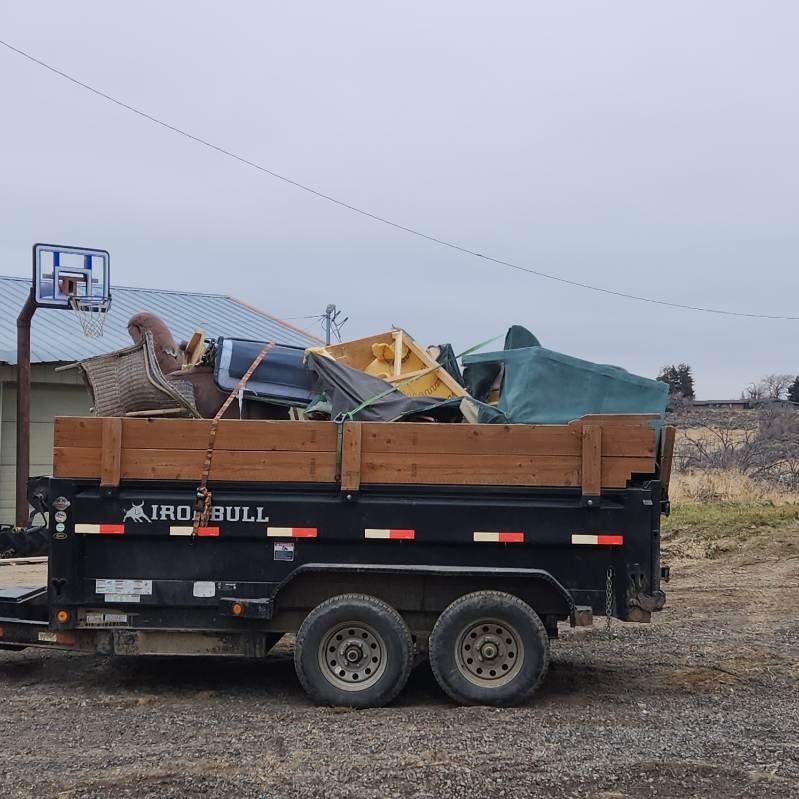 Black dump trailer loaded with assorted scrap metal and debris in a gravel lot under a gray sky