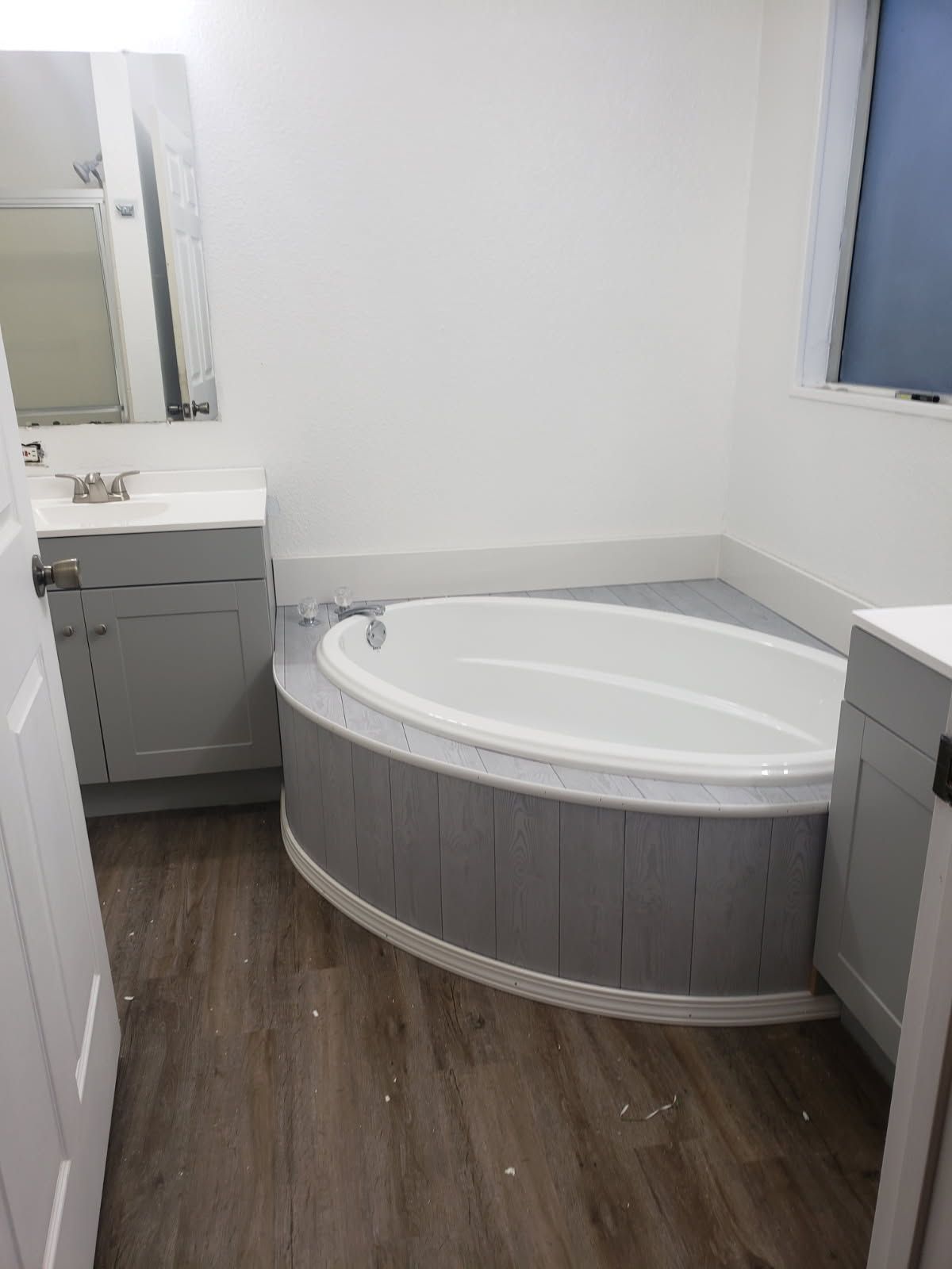 Small bathroom with a white oval bathtub, gray vanity, and wood-look floor.