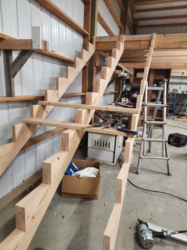 Partially built wooden staircase in a workshop with tools, ladders, and lumber around it