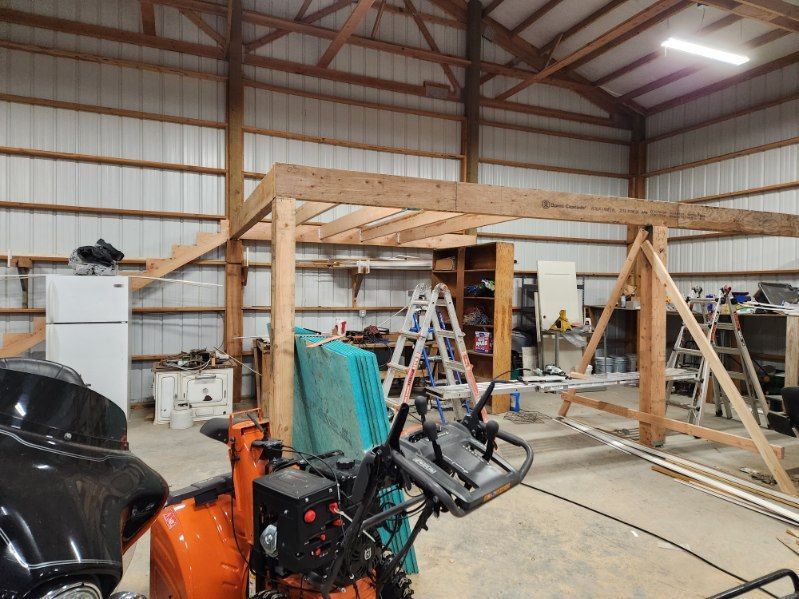 Wood-framed structure under construction inside a metal workshop, with ladders, tools, and equipment around it.