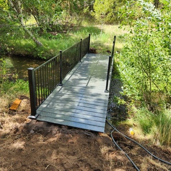 Small metal footbridge over a creek in a wooded area, with black railings and a gray deck.