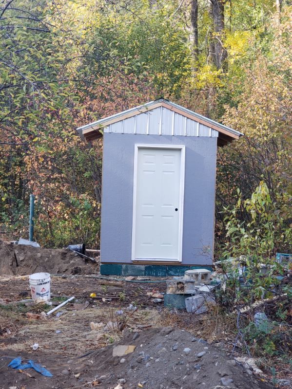 Small gray shed with white door in a wooded yard, surrounded by dirt and debris.