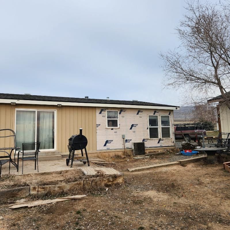Damaged beige house exterior with broken siding, a grill, patio chairs, and leafless tree in yard