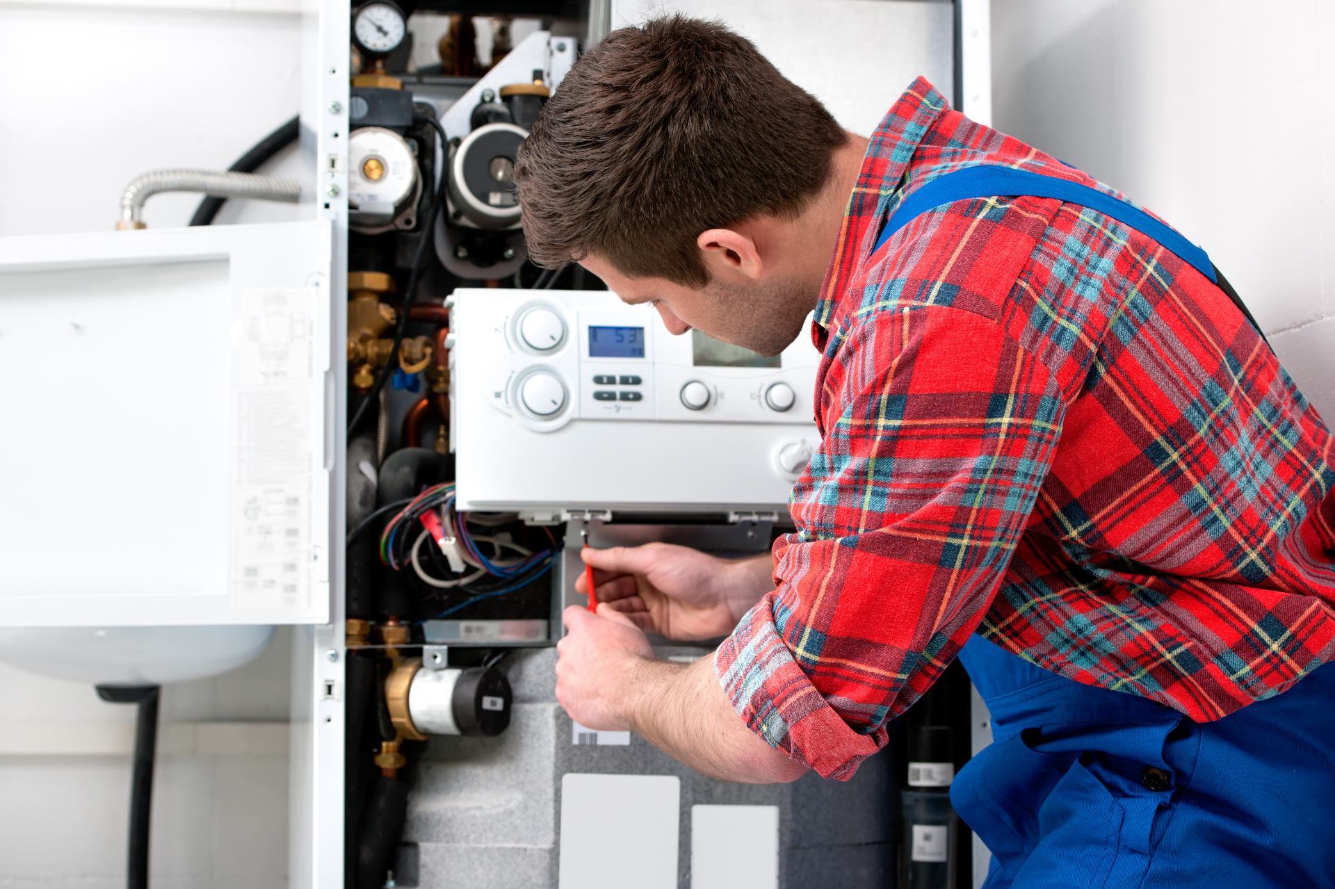 Technician in red plaid shirt repairs a wall-mounted boiler with the cover open.