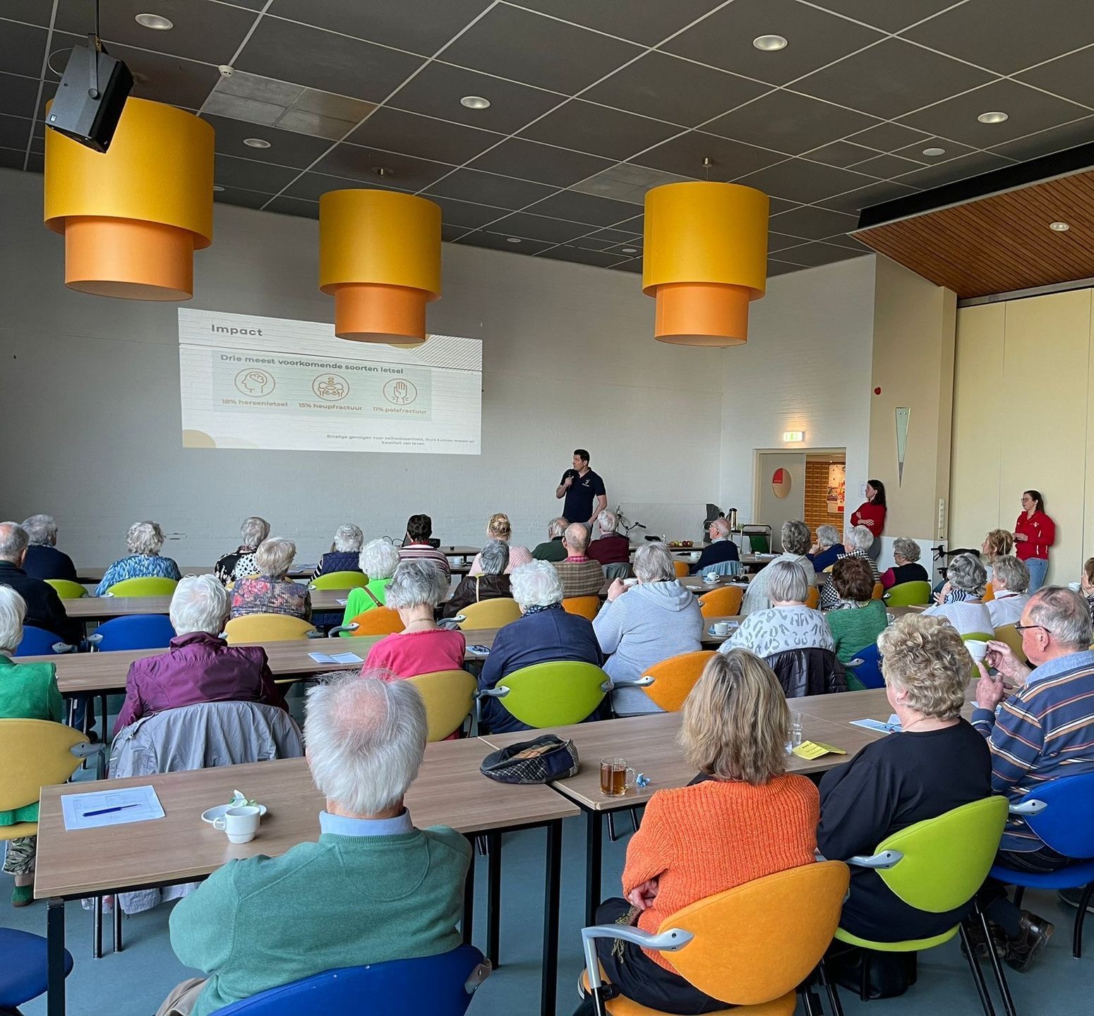 Een presentatie in een grote zaal, het publiek zit aan tafels tegenover een projectiescherm. Drie gele en oranje lampen hangen aan het plafond.