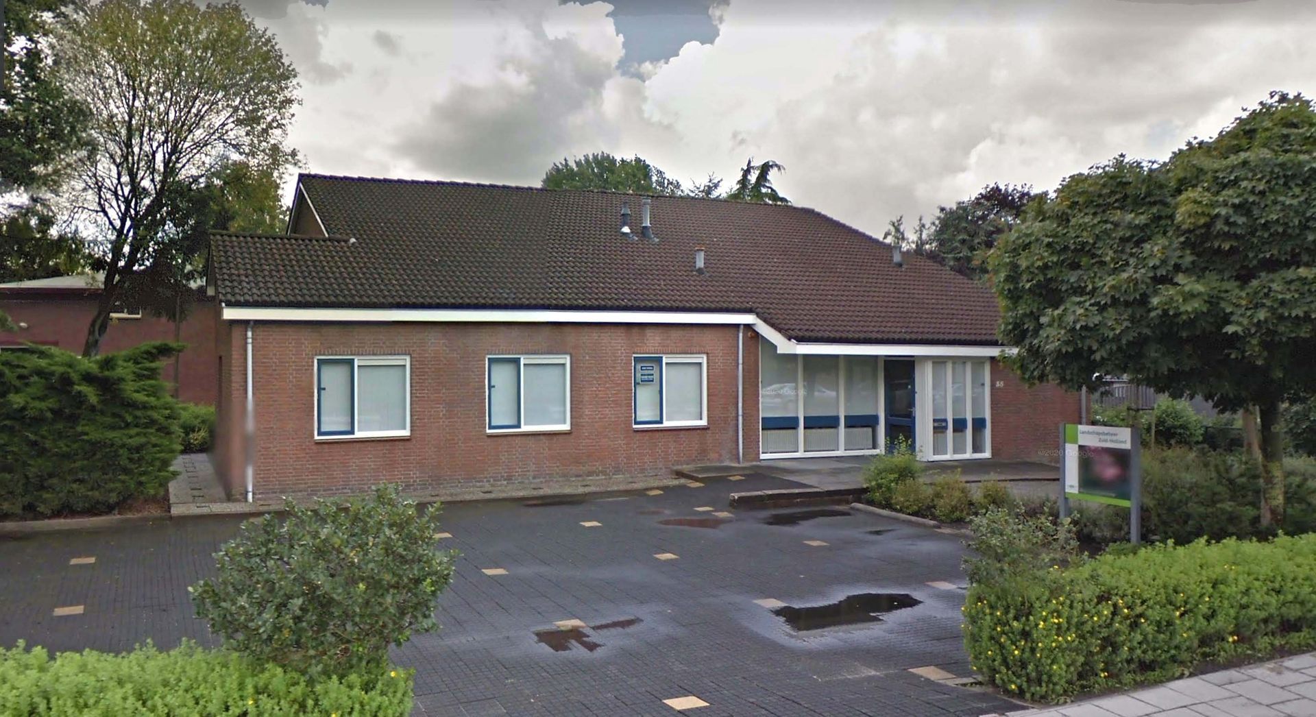 Een bakstenen gebouw met meerdere ramen, omgeven door bomen en een verharde parkeerplaats.