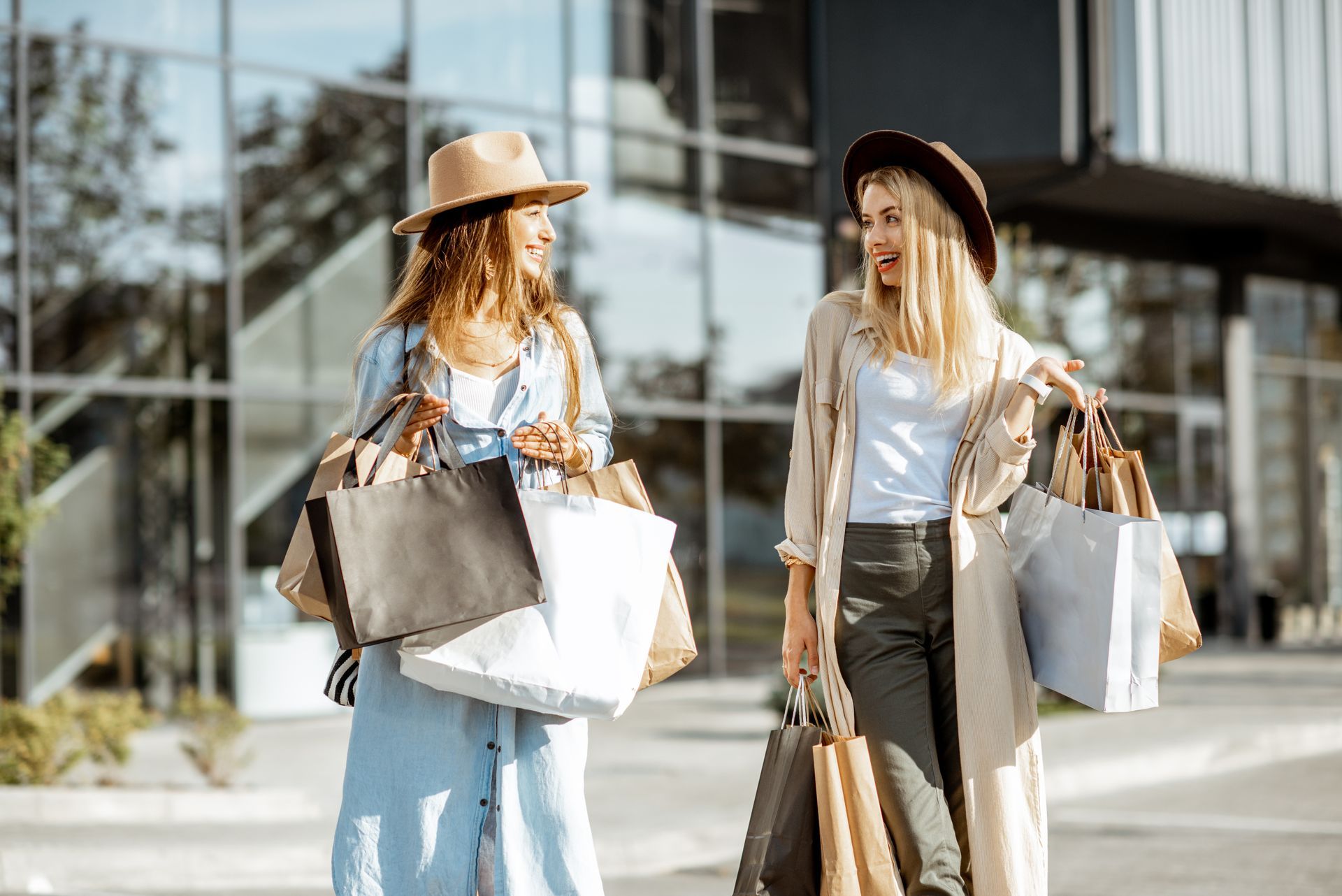 Alt Text: Two smiling young women wearing wide-brimmed hats walk side-by-side outside, carrying numerous paper shopping bags. They are looking at each other and chatting happily in front of a modern building with a large glass facade on a sunny day.