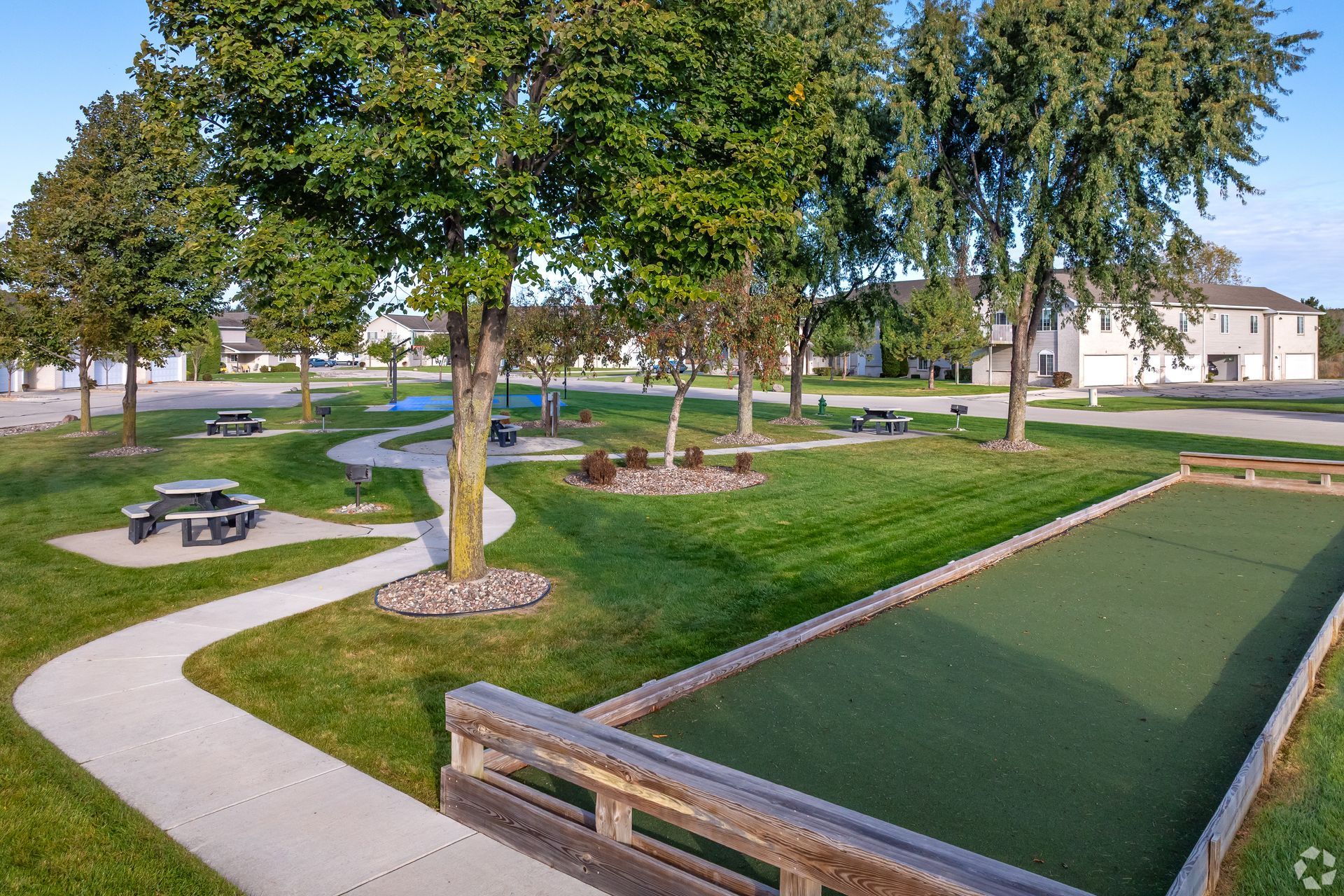 Park with curved paths, trees, picnic tables, and a green putting area beside houses.