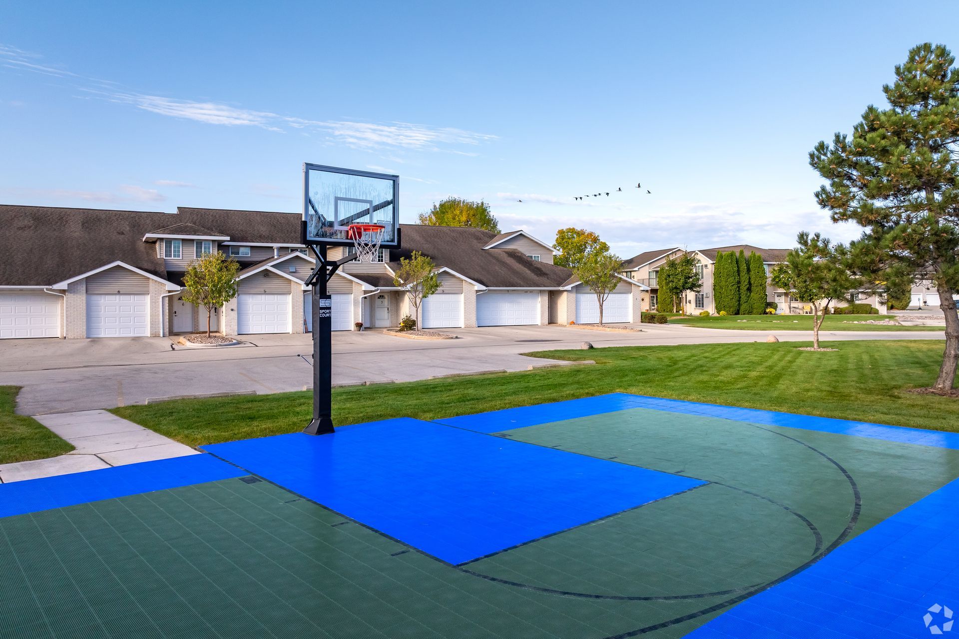 Outdoor basketball court with blue and green surface beside suburban houses under a clear sky