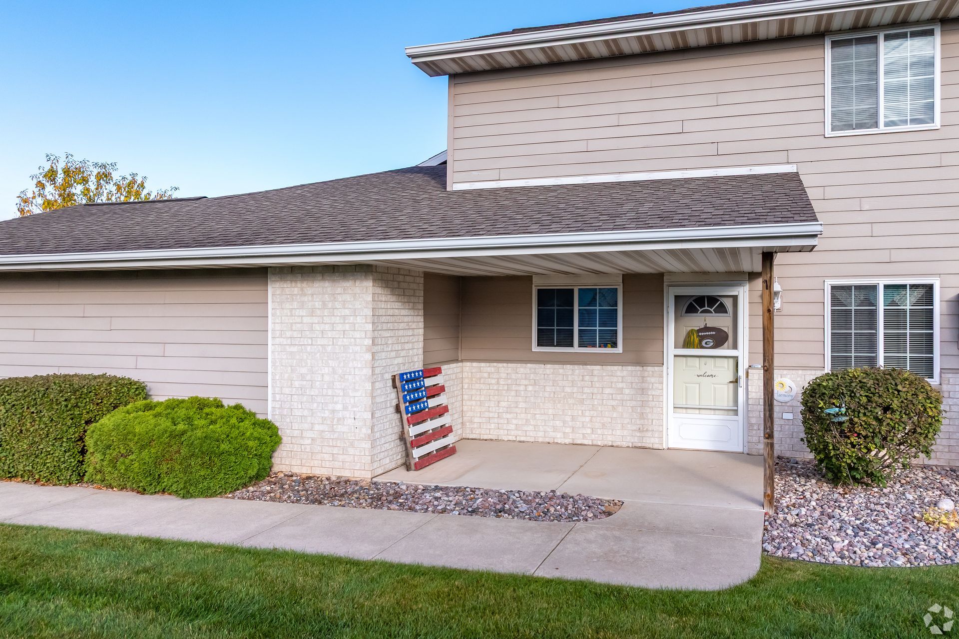 Front porch of a beige townhouse with white door, trimmed shrubs, and an American flag sign.