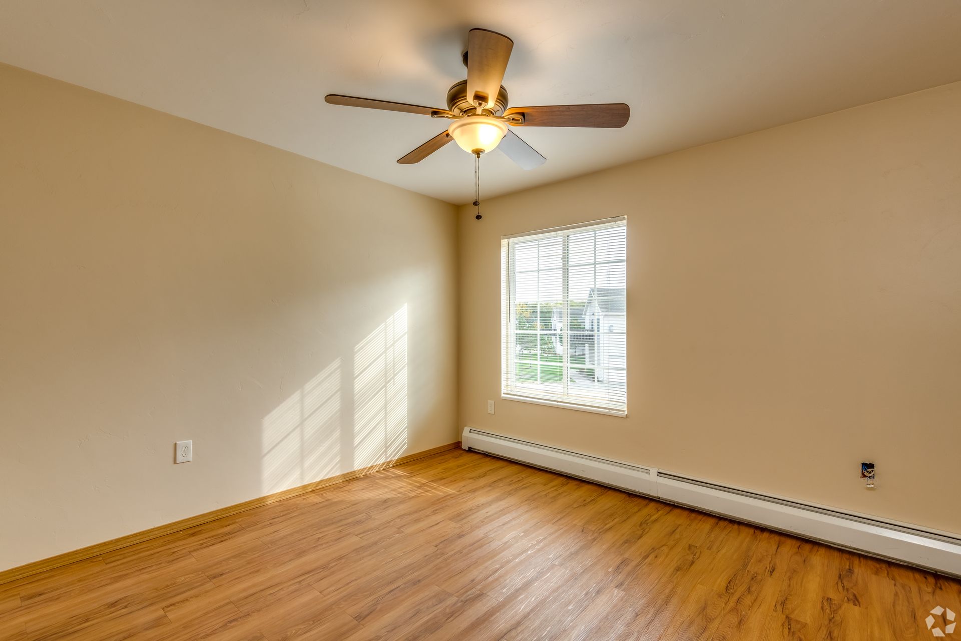 Empty beige room with hardwood floor, ceiling fan, and sunlight through a window