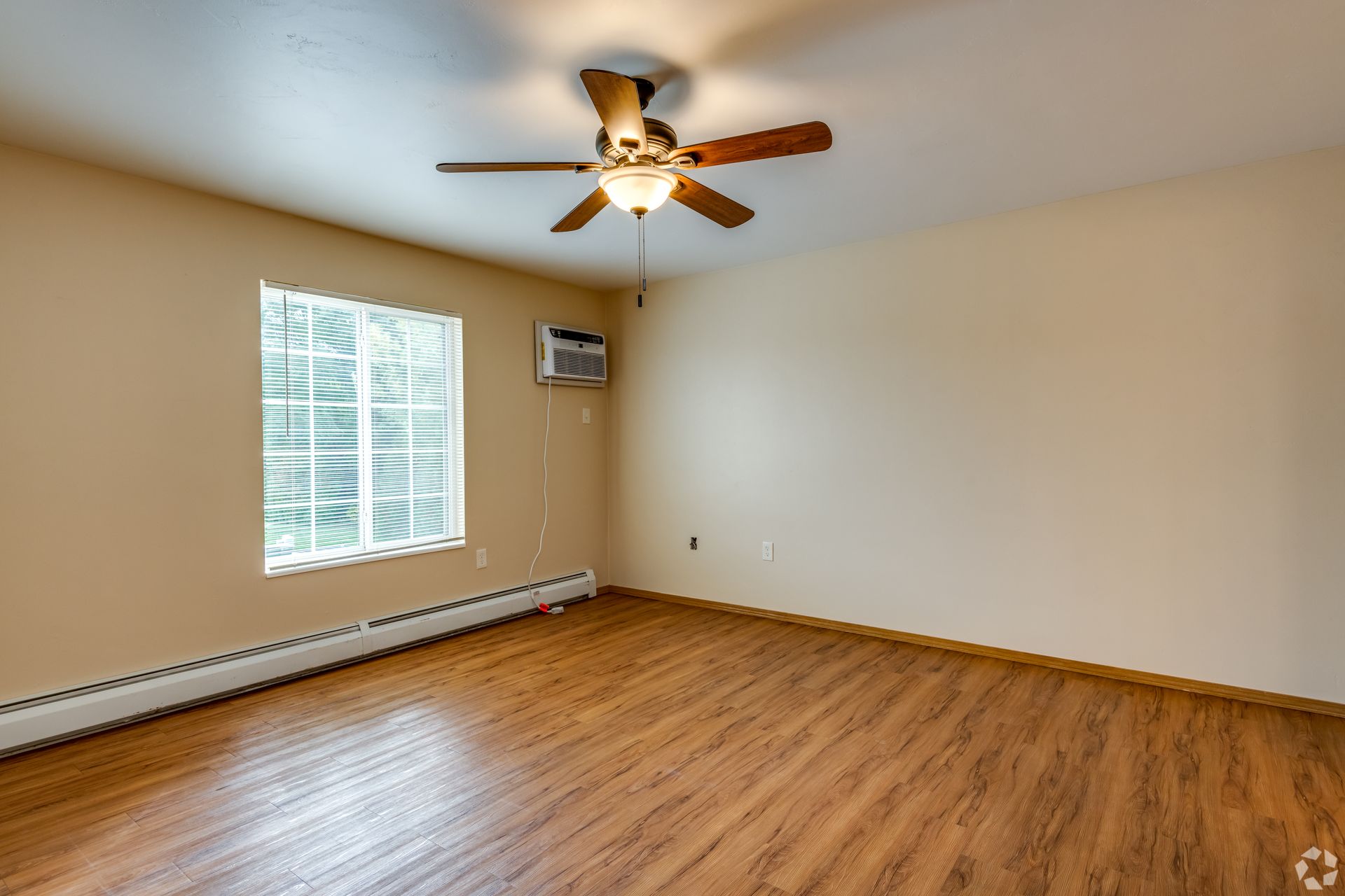 Empty beige room with wood floors, a ceiling fan, window blinds, and a wall heater.