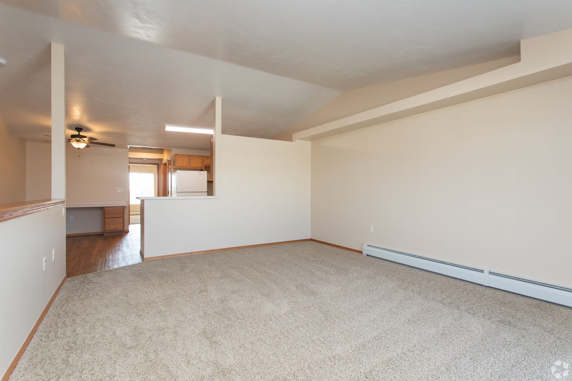 Empty carpeted living room with white walls and a half wall opening to the kitchen area