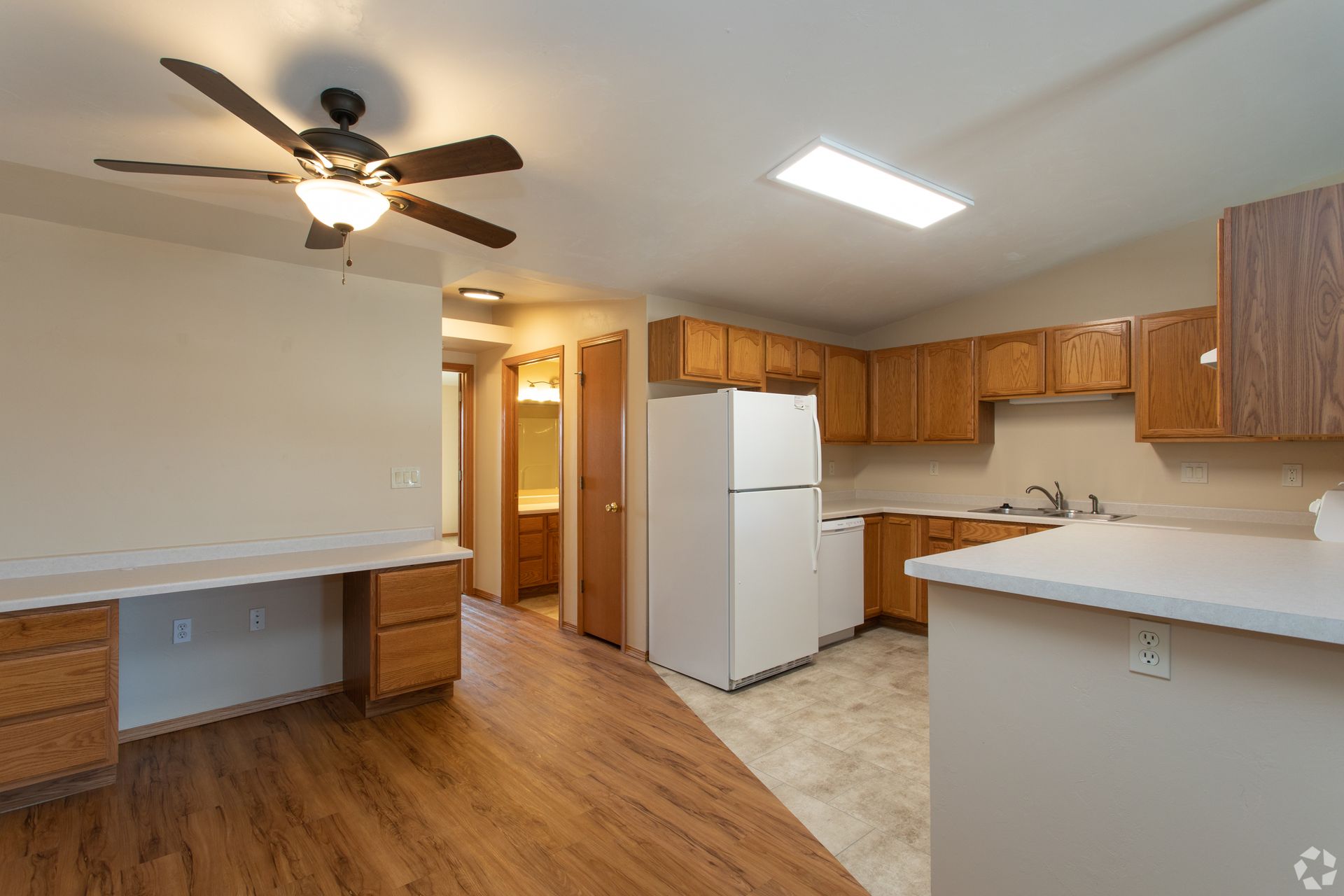 Empty kitchen with wood floors, white appliances, brown cabinets, and a ceiling fan by a small dining area