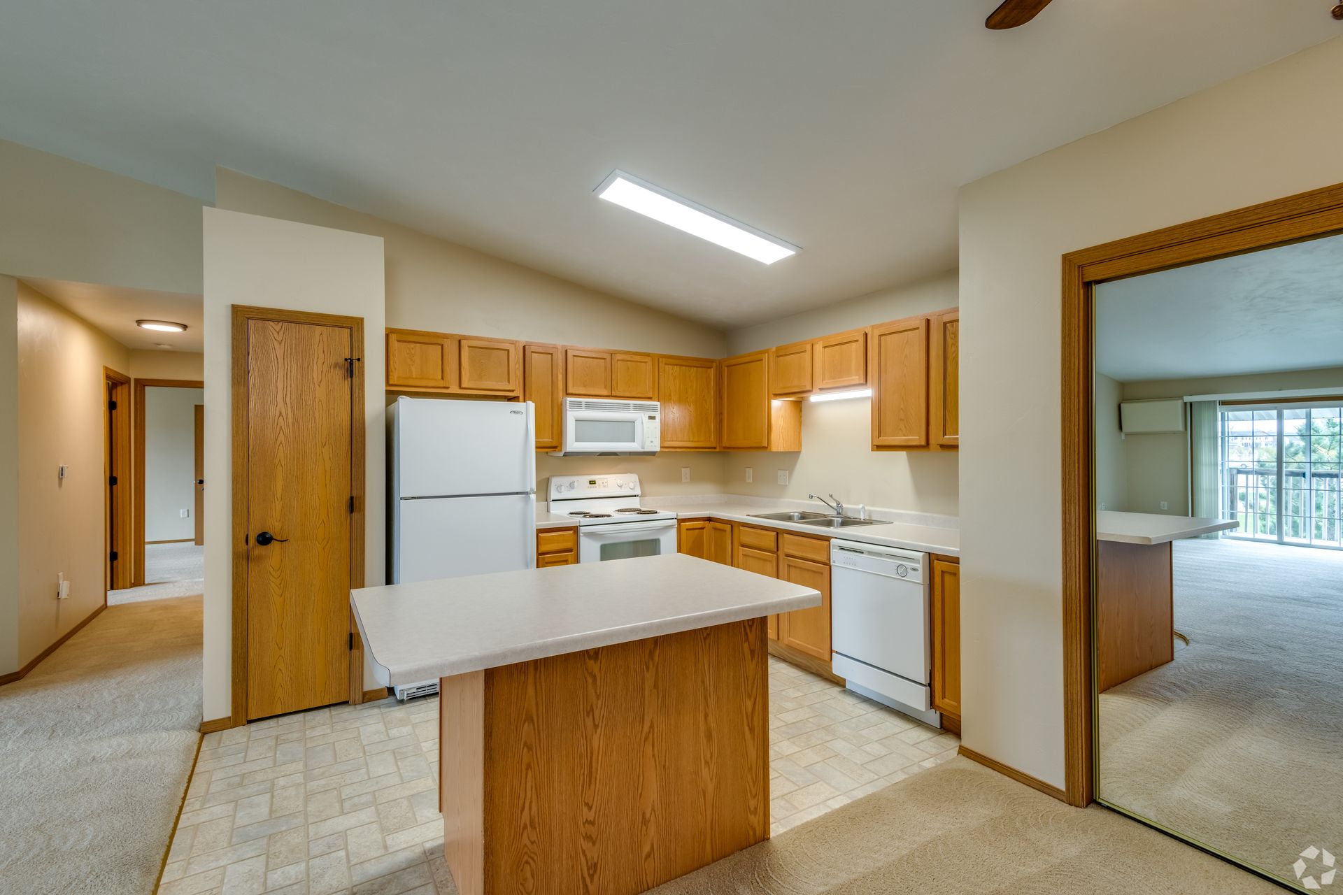 Open-plan kitchen with white appliances, wood cabinets, and carpeted adjacent rooms