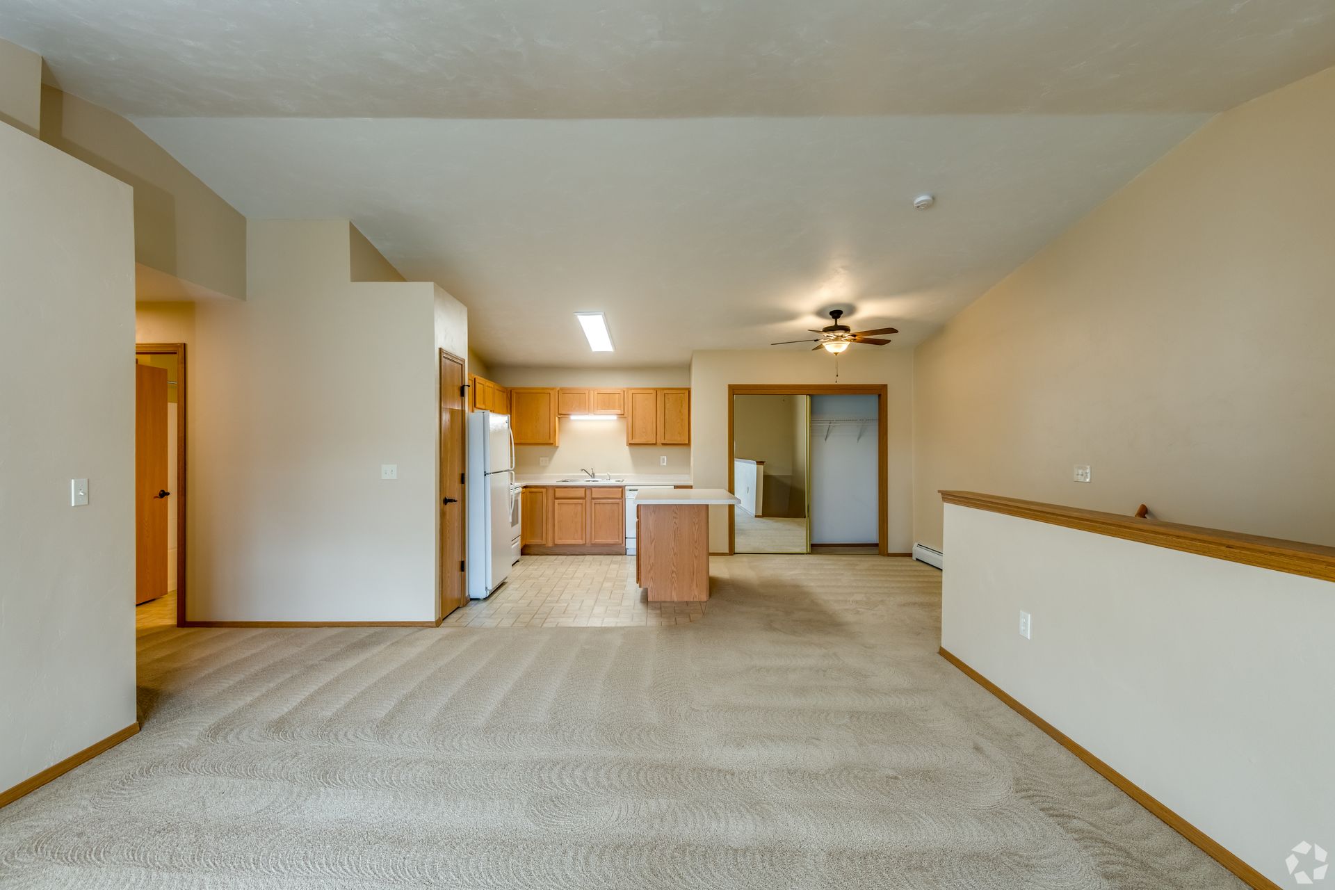 Empty carpeted living room with open kitchen, beige walls, and a breakfast bar.