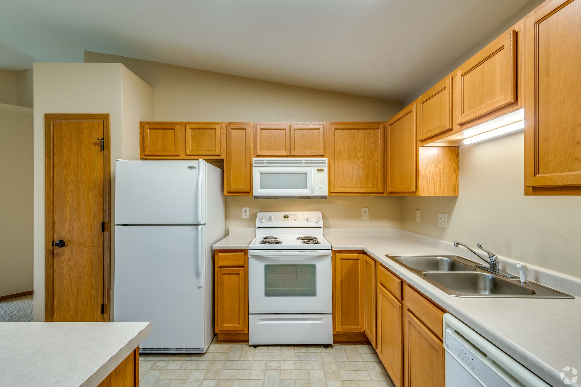 Small kitchen with wood cabinets, white appliances, and a sink beside a countertop.